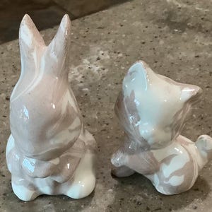 May include: Two ceramic figurines featuring a glossy, marbled finish in shades of cream and taupe. One figurine is shaped like a rabbit with long, upright ears, while the other is shaped like a small cat with pointed ears and a curled tail.