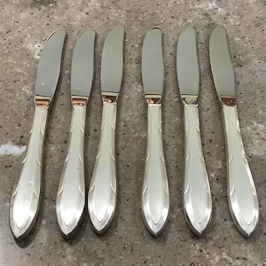 May include: Six silver butter knives with curved blades and decorative handles. The knives are arranged in a row, showcasing their polished finish and elegant design. The handles have a subtle, elongated shape.