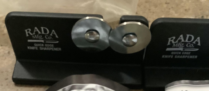 May include: Black knife sharpener with two silver sharpening wheels. The text "RADA Mfg. Co. QUICK EDGE KNIFE SHARPENER" is printed in white on the black base. The sharpener is designed for quick and easy knife sharpening.