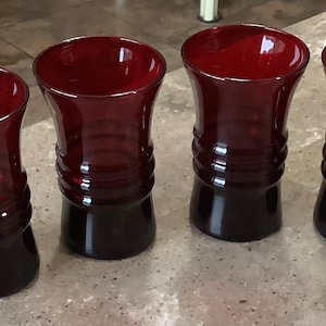 May include: Four ruby red glass tumblers with a flared top and a dark base. The glasses have a ribbed design around the middle. The tumblers are arranged in a row and are suitable for serving beverages.
