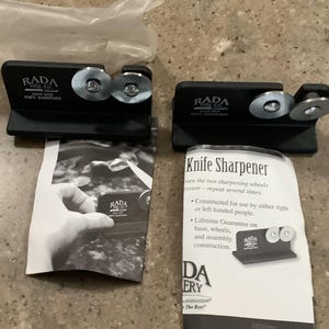 May include: Two black knife sharpeners with silver sharpening wheels. Each sharpener has the text "RADA Quick Edge Knife Sharpener". The sharpeners are on top of instruction manuals with the text "Knife Sharpener".
