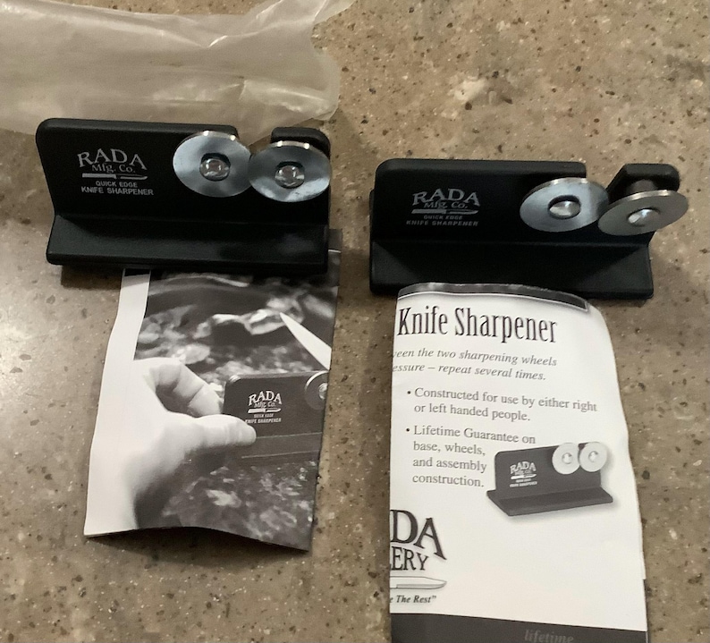 May include: Two black knife sharpeners with silver sharpening wheels. Each sharpener has the text "RADA Quick Edge Knife Sharpener". The sharpeners are on top of instruction manuals with the text "Knife Sharpener".