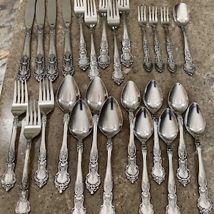 May include: A collection of ornate silverware, including knives, forks, and spoons, arranged on a neutral surface. The silverware features detailed floral and scrollwork designs on the handles, with a polished, reflective finish. The set includes various sizes of forks, spoons, and knives.
