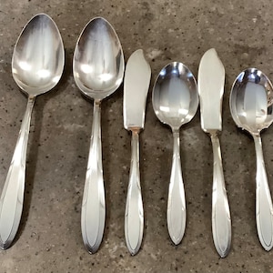 May include: A set of six silver-colored utensils, including two spoons, two butter knives, and two smaller spoons. The utensils have a simple, elegant design with a reflective surface.