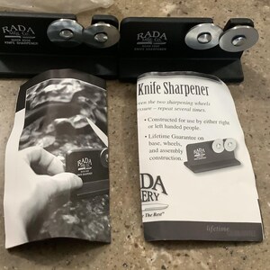 May include: Two black RADA knife sharpeners with silver sharpening wheels. The text "RADA Mfg. Co. QUICK EDGE KNIFE SHARPENER" is printed on the sharpeners. Instruction manuals are also included, with text that reads "Knife Sharpener".