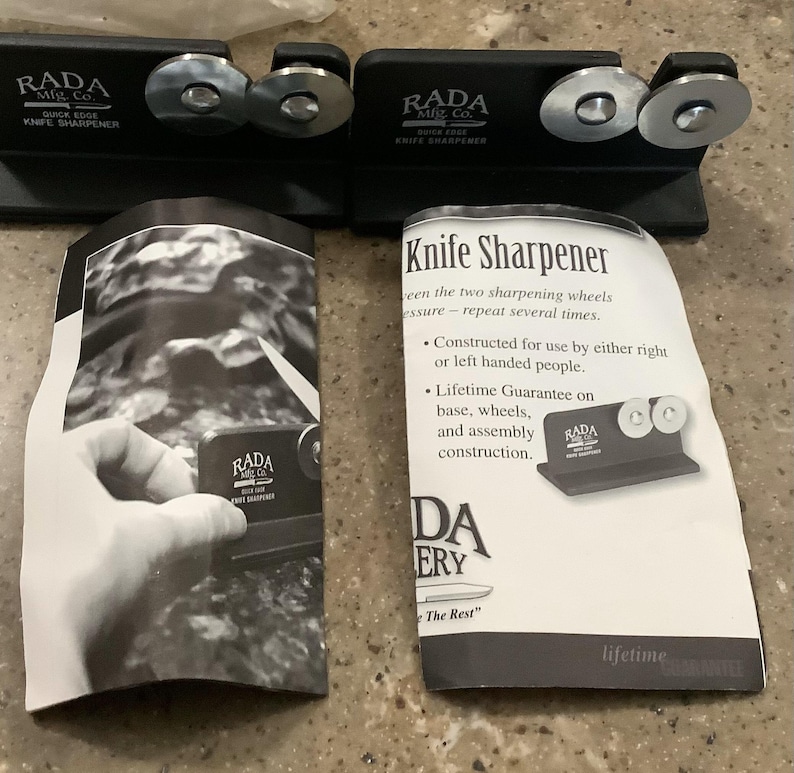 May include: Two black RADA knife sharpeners with silver sharpening wheels. The text "RADA Mfg. Co. QUICK EDGE KNIFE SHARPENER" is printed on the sharpeners. Instruction manuals are also included, with text that reads "Knife Sharpener".