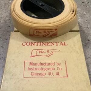May include: A beige paper tape roll with a black plastic center spool, resting on a matching beige cardboard box. The tape and box feature red ink stamps of a pointing finger icon with the text No. 6, CONTINENTAL, and Manufactured by Instructograph Co. Chicago 40, Ill.