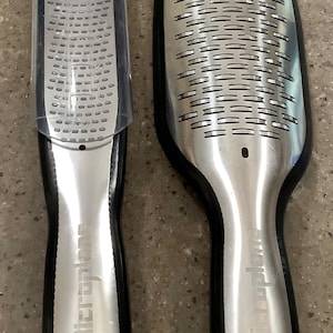May include: Two Microplane graters with black handles and stainless steel grating surfaces. One grater is rectangular with a clear plastic cover, and the other is paddle-shaped. The brand name is printed on the handles.