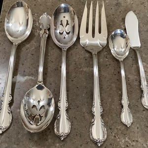May include: A collection of ornate, silver-colored silverware, including spoons, a fork, and a butter knife. The silverware has detailed floral patterns on the handles. The spoons include a serving spoon, a slotted spoon, and a soup spoon.