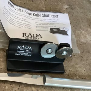 May include: A black RADA Quick Edge Knife Sharpener with two sharpening wheels and a stainless steel knife. The packaging includes the text "R119 Quick Edge Knife Sharpener" and "RADA CUTLERY".