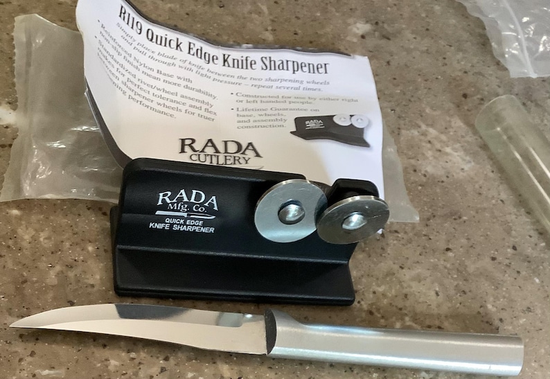 May include: A black RADA Quick Edge Knife Sharpener with two sharpening wheels and a stainless steel knife. The packaging includes the text "R119 Quick Edge Knife Sharpener" and "RADA CUTLERY".