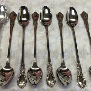 May include: A set of eight silver-toned stainless steel iced tea spoons with long, slender handles and decorative embossed floral detailing at the base of the handle. One spoon is enclosed in a clear plastic sleeve, while seven are unwrapped.
