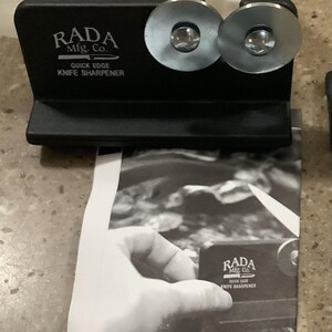May include: Black knife sharpener with two silver sharpening wheels. The text "RADA Mfg. Co. Quick Edge Knife Sharpener" is printed on the front. A black and white image of a hand holding a knife sharpener is in the background.