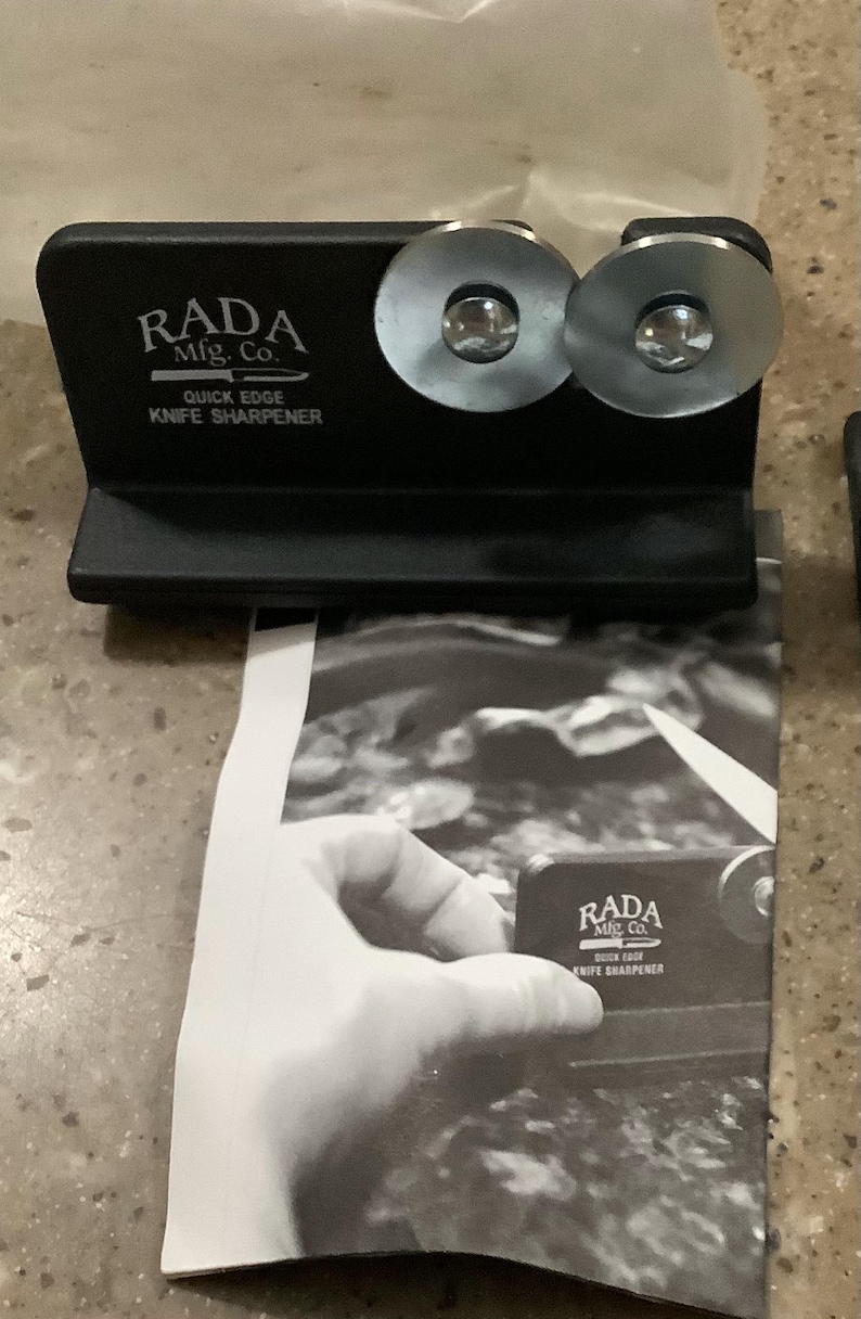 May include: Black knife sharpener with two silver sharpening wheels. The text "RADA Mfg. Co. Quick Edge Knife Sharpener" is printed on the front. A black and white image of a hand holding a knife sharpener is in the background.