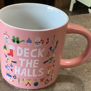 May include: A light pink ceramic mug with a white interior and a handle on the right side. The exterior features the white text DECK THE HALLS surrounded by colorful illustrations of holiday ornaments, a bird, a boot, and festive icons.