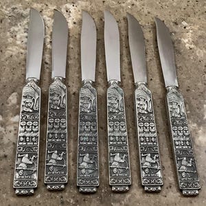 May include: Set of six silver butter knives. Each knife features a serrated blade and an ornate handle with engraved details, including figures and text. The handles have the text "HEILAG BLAV" and "06 BARS MENW".
