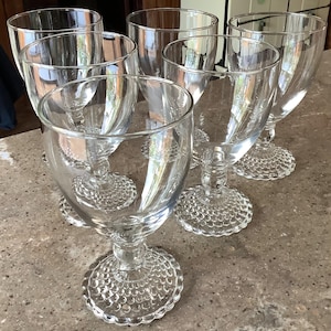 May include: Set of six clear glass goblets. Each goblet features a wide bowl, a stem, and a textured, circular base. The glasses are arranged on a neutral-toned surface, reflecting light and creating a sense of elegance.