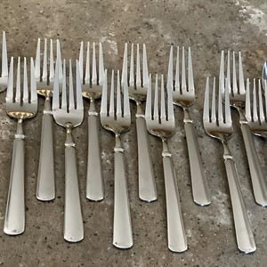 May include: A collection of eleven stainless steel dinner forks with four tines and a smooth, flat handle design, arranged alongside a single slotted serving spoon and a portion of another spoon, all featuring a polished, reflective metallic finish.