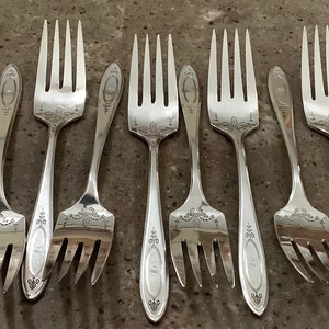 May include: A collection of silver forks with ornate floral and scrollwork designs. The forks are arranged on a gray surface, showcasing their polished finish and elegant details. Ideal for formal dining.