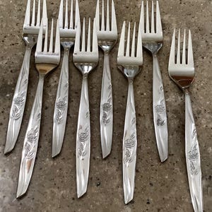 May include: A collection of nine silver forks with a floral rose design on the handles. The forks are arranged on a neutral-toned surface. The tines are evenly spaced, and the handles have a detailed embossed pattern.