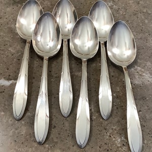 May include: Seven silver spoons arranged on a gray surface. The spoons have an oval bowl and a long, slender handle with a decorative design. The spoons are reflective, showing the surrounding environment.