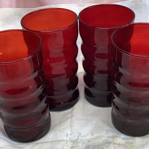 May include: Four red-colored drinking glasses. Each glass has a cylindrical shape with a series of horizontal, rounded ridges. The glasses are made of a transparent material, allowing light to pass through, and are set on a white surface.