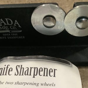 May include: Black knife sharpener with two silver sharpening wheels. The text "RADA Mfg. Co. QUICK EDGE KNIFE SHARPENER" is printed on the sharpener. A white paper with the text "Knife Sharpener" is in the foreground.