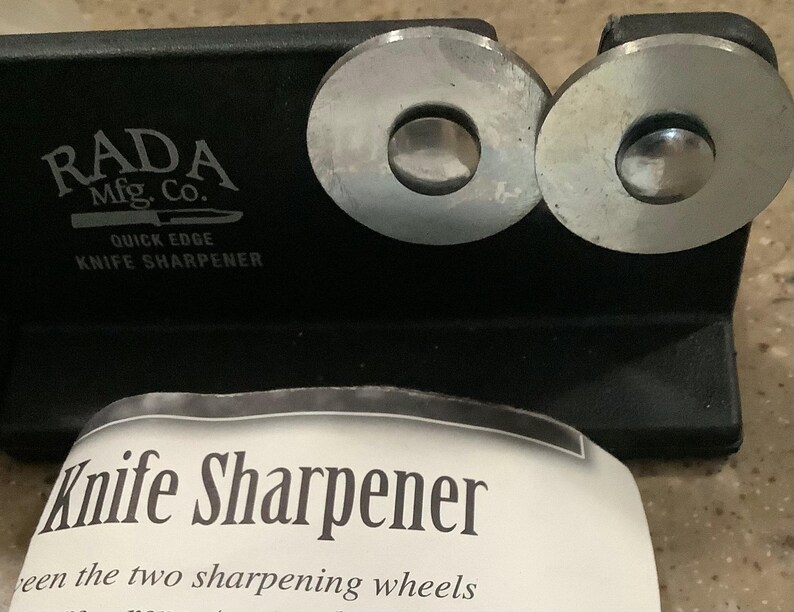 May include: Black knife sharpener with two silver sharpening wheels. The text "RADA Mfg. Co. QUICK EDGE KNIFE SHARPENER" is printed on the sharpener. A white paper with the text "Knife Sharpener" is in the foreground.
