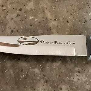 May include: A stainless steel knife with a silver handle. The blade has a curved edge and features the text "Donovan Farmers Co-op" along with a logo. The knife is designed for kitchen use.