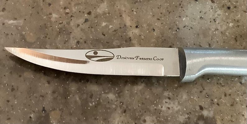May include: A stainless steel knife with a silver handle. The blade has a curved edge and features the text "Donovan Farmers Co-op" along with a logo. The knife is designed for kitchen use.