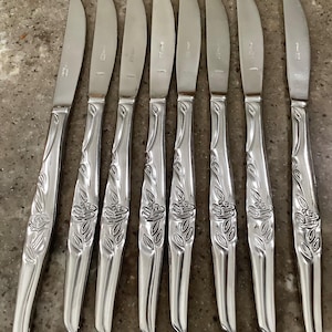 May include: A set of eight stainless steel dinner knives. Each knife features a floral design on the handle and a serrated edge. The knives are arranged on a neutral-toned surface.