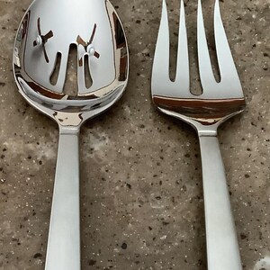 May include: A shiny, stainless steel serving spoon with slotted design and a matching serving fork. The spoon has a rounded bowl with cut-out details. The fork has four tines. Both utensils have long, flat handles. Kitchen and dining.