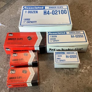 May include: A collection of six rectangular cardboard boxes containing binder clips. The boxes are labeled Associated or CLI, with text indicating sizes such as 1 inch, 5/8 inch, and 3/8 inch capacity, and quantities of one dozen per box.