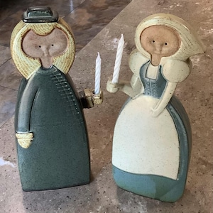 May include: Two ceramic candle holders shaped like stylized figures. The left figure wears a dark green coat and hat. The right figure wears a blue and white outfit with a head covering. Each figure holds a thin, white, spiral-patterned taper candle.