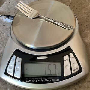 May include: A silver-toned metal fork with an intricate floral pattern on the handle, resting on a digital kitchen scale. The scale display shows a weight of 15.7 grams, with a maximum capacity of 1000 grams, 36 ounces, or 0.035 ounces.