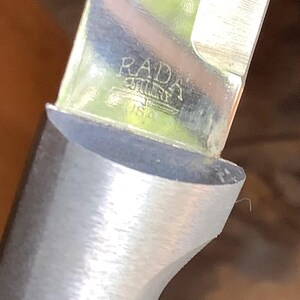 May include: Close-up of a knife blade with a silver handle. The blade is shiny and reflects the surrounding environment. The text "RADA" and "USA" are visible on the blade. The knife appears to be new and unused.