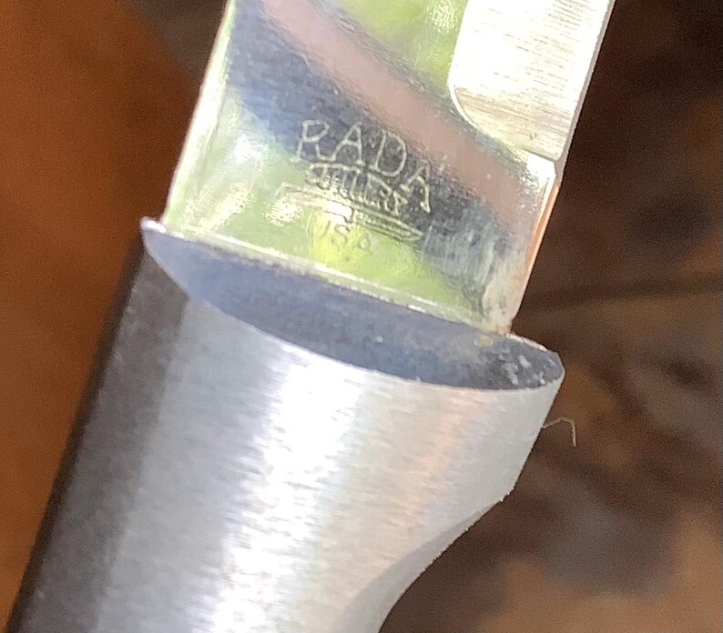 May include: Close-up of a knife blade with a silver handle. The blade is shiny and reflects the surrounding environment. The text "RADA" and "USA" are visible on the blade. The knife appears to be new and unused.