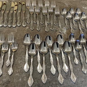 May include: An assortment of silver-colored flatware, including knives, forks, and spoons, arranged on a gray surface. The silverware has ornate, decorative handles. The spoons have a rounded bowl shape. The knives have a smooth, shiny blade.