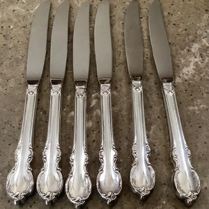 May include: Six silver-colored knives with ornate handles are arranged on a speckled surface. The knives have a classic design, with a smooth, curved blade and a decorative handle featuring floral-like patterns.