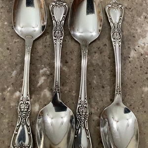 May include: Four silver-toned metal spoons arranged in a row. Each spoon features an ornate, embossed floral pattern along the handle, extending from the base of the bowl to the rounded tip of the handle, with a smooth, polished finish.