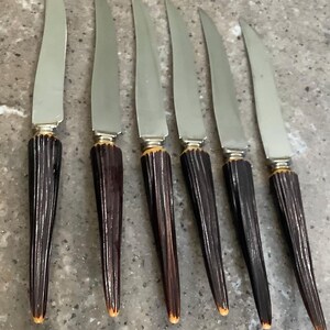 May include: Six vintage steak knives with stainless steel blades and dark brown, ribbed handles. The handles have a gold-colored accent near the blade. The knives are arranged in a row, showcasing their design and craftsmanship.
