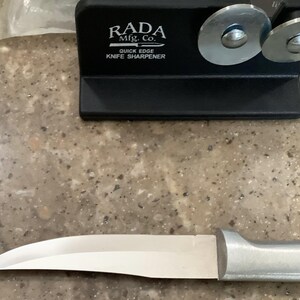 May include: A black knife sharpener with the text "RADA Mfg. Co. QUICK EDGE KNIFE SHARPENER" and two silver sharpening wheels. A silver-bladed knife with a silver handle is in the foreground. The knife is designed for kitchen use.