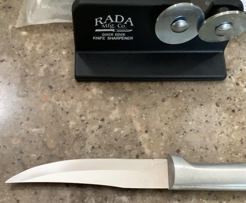 May include: A black knife sharpener with the text "RADA Mfg. Co. QUICK EDGE KNIFE SHARPENER" and two silver sharpening wheels. A silver-bladed knife with a silver handle is in the foreground. The knife is designed for kitchen use.