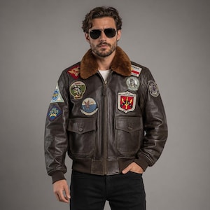 Könnte beinhalten: Eine dunkelbraune Leder-Bomberjacke mit braunem Kunstfellkragen, durchgehendem Frontreißverschluss und zwei Fronttaschen mit Klappen. Die Jacke ist mit verschiedenen gestickten militärischen Aufnähern und dem Text U.S.S. WILLIAM H. STANDLEY verziert.