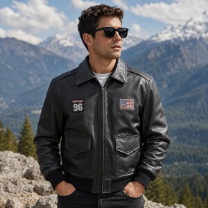 Könnte beinhalten: Eine schwarze Leder-Bomberjacke mit Frontreißverschluss, spitzem Kragen und gerippten Bündchen. Die Jacke hat einen Aufnäher auf der rechten Brust mit dem Text VINTAGE 96 und einen Aufnäher auf der linken Brust mit einer amerikanischen Flagge.