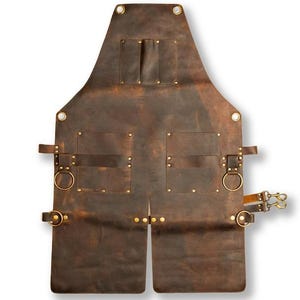 Könnte beinhalten: Eine braune Lederschürze mit mehreren Taschen und Messingbeschlägen. Die Schürze hat verstellbare Riemen, Metallringe und ein geteiltes Beingestaltung. Sie verfügt über eine robuste, abgenutzte Oberfläche, die für verschiedene Bastel- oder Werkstattaktivitäten geeignet ist.
