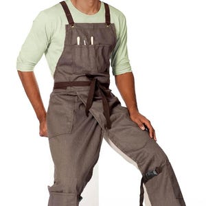 May include: Brown apron with matching pants. The apron has a chest pocket with two utensils. The pants have a knee strap. The apron has brown straps and a waist tie. The person is wearing a light green long-sleeve shirt.