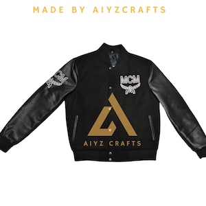Puede incluir: Chaqueta universitaria negra con mangas de cuero. La chaqueta presenta las letras "MCM" y "AIYZ CRAFTS" en blanco y dorado. La chaqueta tiene una botonadura y cuello y puños acanalados.