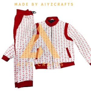 Op de afbeelding: Een witte trainingspak met rode accenten en een herhalend patroon. De jas heeft een ritssluiting en zakken. De bijpassende broek heeft een taille met trekkoord. De tekst "MADE BY AIYZCRAFTS" staat bovenaan.