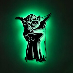 Könnte beinhalten: Eine LED-Wandleuchte in Form der Figur Yoda, die ein schwarzes Silhouettendesign aufweist und ein helles grünes Leuchten von den Kanten ausstrahlt, wodurch ein deutlicher Halo-Effekt auf der umgebenden Wandfläche entsteht.